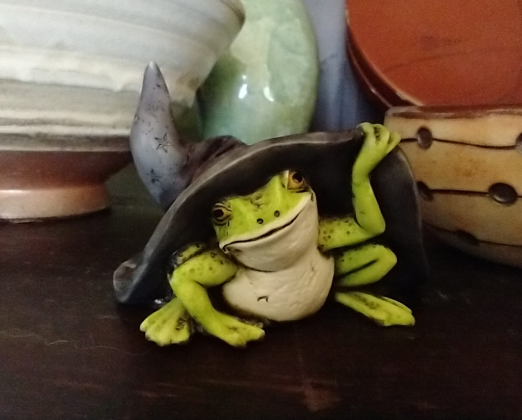 Ceramic frog looking out from under a witch hat.