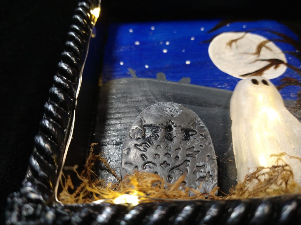 Closeup of the corner of a shadowbox with a lit ghost and a fancy tombstone.