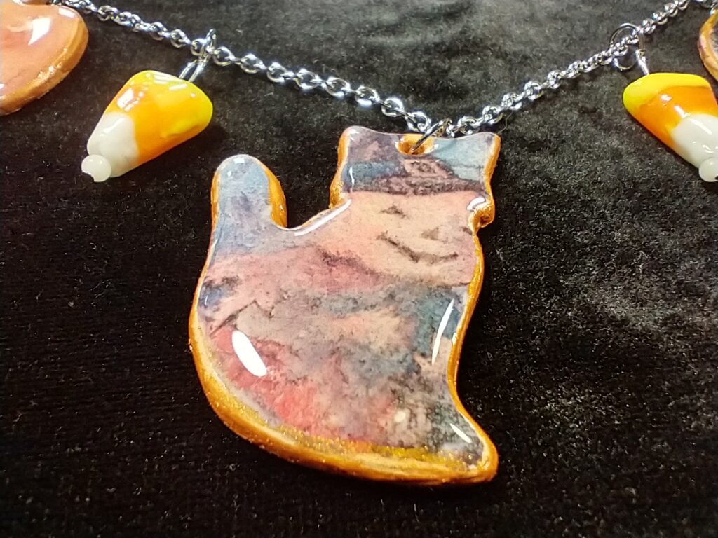 Closeup of cat-shaped charm with a jack-o-lantern witch on it.  There are candy corn beads on either side.