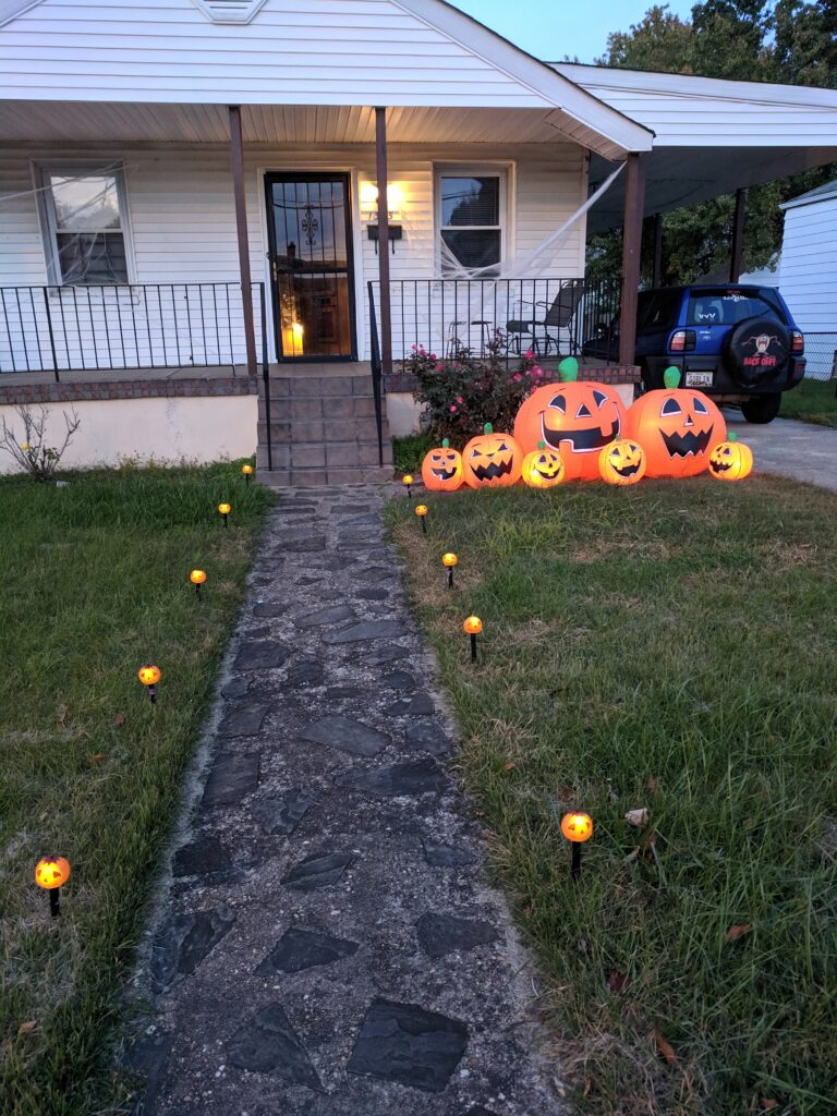 A path lined with little orange lights leads to a small white house.  To the left of the path is a large inflatable of a family of jack-o-lanterns.