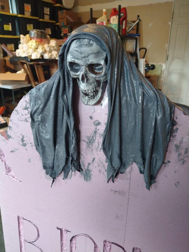 A pink foam tombstone topped with a skull that has been draped with a hood soaked with monster mud.