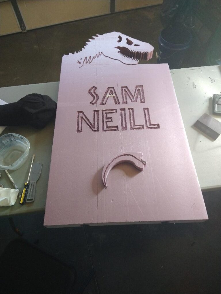 A pink foam tombstone for Sam Neill, with the top carved in a T-Rex skull, and a raptor claw at the bottom.