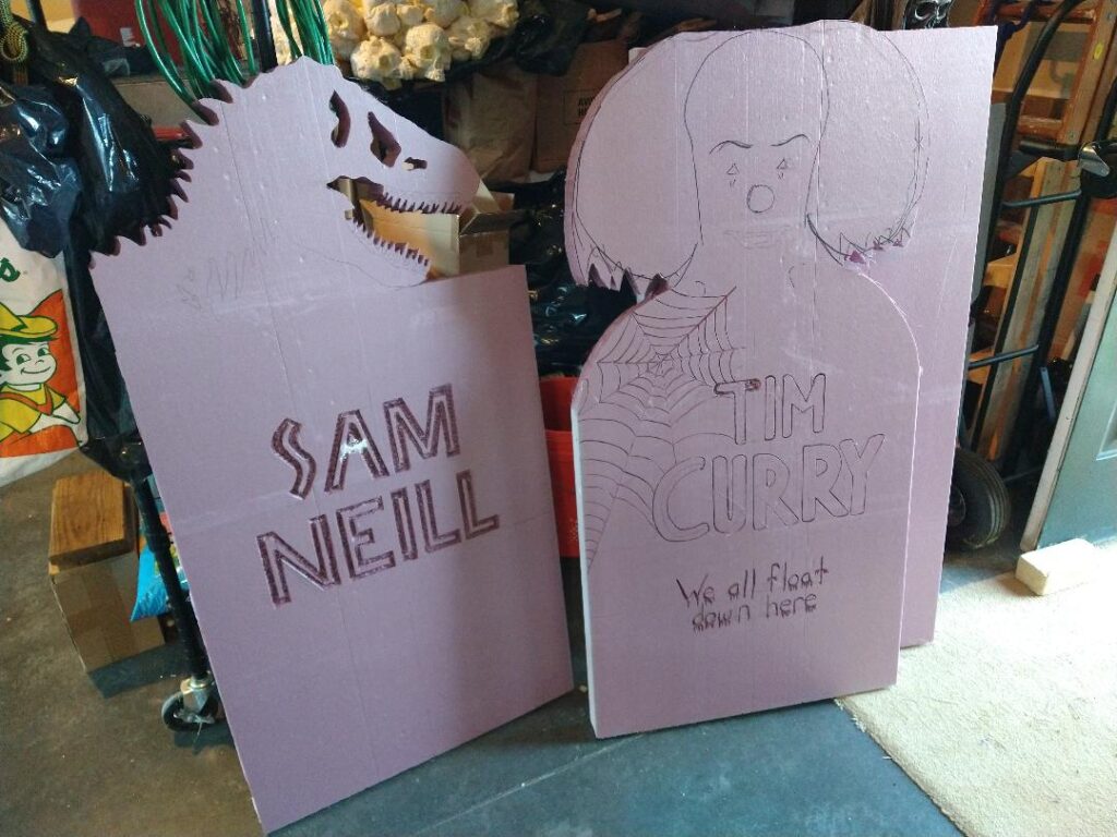 Left is a pink foam tombstone with a t-rex skull, and the name SAM NEILL. Right is a clown head and TIM CURRY with a spiderweb coming off the name. Below it says "We all float down here."